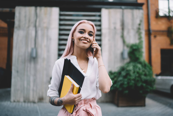 Cheerful Caucasian schooler with education notepad and sketchbook for design using smartphone application for making positive international call, happy pink haired student enjoying mobile talk - Fotografie, Obrázek