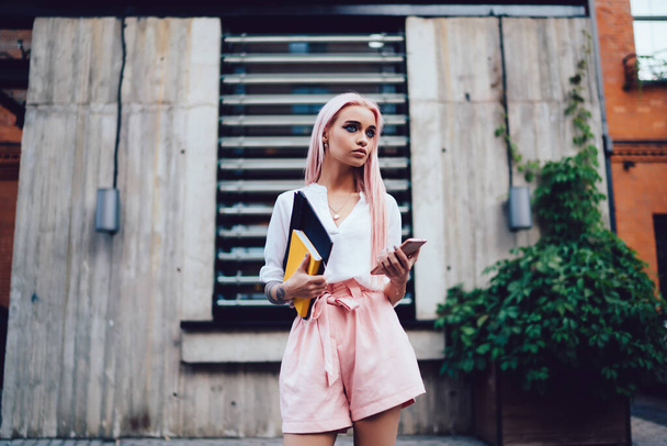 Pensive pink haired female student with education notebooks and modern cellphone device standing at city urban setting and thoughtful looking away, contemplative designer pondering on mobile update - 写真・画像