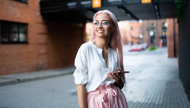 Cheerful Caucasian blogger holding modern cellphone device smiling while remembering good times, carefree millennial teenager in spectacles for provide eyes protection feeling happy at urban setting - Φωτογραφία, εικόνα