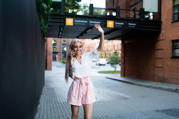 Happy Caucasian hipster girl in trendy eyewear for vision protection standing at city urbanity and waving while waiting best friend, cheerful millennial calling via smartphone with roaming connection - 写真・画像