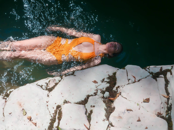 Una chica en un traje de baño amarillo acostada sobre su espalda nada en el agua cerca de la costa rocosa - Foto, Imagen