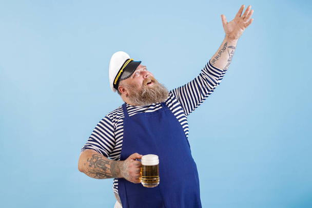 Позитивный толстяк в матросском костюме держит пиво, поднимая руку на синем фоне. - Фото, изображение