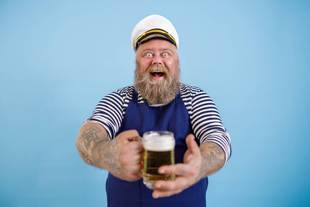 Glücklicher dicker Mann im Matrosenanzug bietet einen Becher frisches Bier auf hellblauem Hintergrund an - Foto, Bild