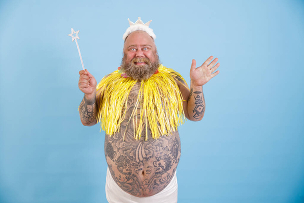 Vidám szakállas kövér ember korona, mágikus bot és sárga köpeny világoskék háttér - Fotó, kép
