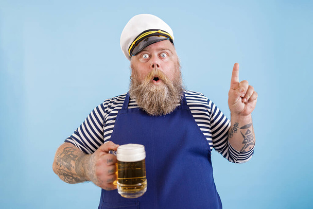 Συναισθηματικός ναύτης με υπέρβαρο κρατά μπύρα και χειρονομίες σε γαλάζιο φόντο - Φωτογραφία, εικόνα