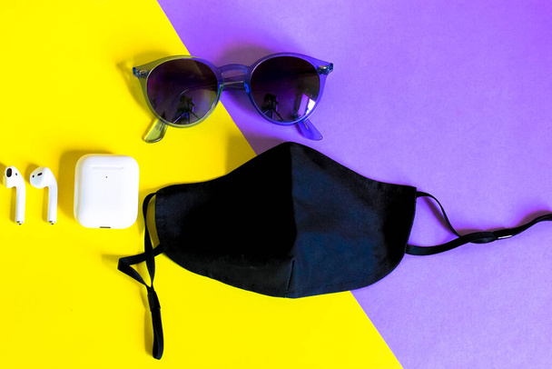黒い医療用フェイスマスク、紫色のサングラス、黄色紫色の斜めの背景にワイヤレスヘッドフォン付きのイヤフォンをフラット。新しい正常よ。Pandemic Covid 19.スタイリッシュなアクセサリー夏休みトップビュー. - 写真・画像
