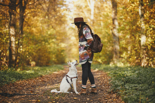 Stylish woman traveler in hat hiking with cute dog in sunny autumn woods. Young female hipster traveling with swiss shepherd white dog. Travel with pet, loyal companion. Wanderlust - Fotoğraf, Görsel