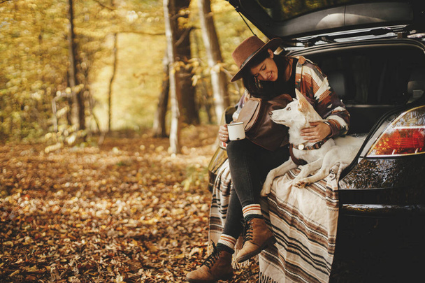 Stylish happy woman traveler sitting with cute dog in car trunk in sunny autumn woods. Travel and road trip with pet. Space for text. Young hipster female travelling with sweet white dog - Photo, Image