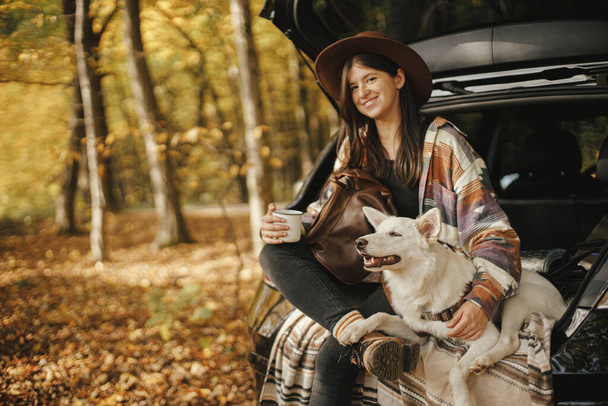 Stílusos hipster nő csésze és hátizsák ül aranyos kutya autó csomagtartóban napos őszi erdőben. Utazás és utazás kedvtelésből tartott állatokkal. Helyet a szövegnek. fiatal női utazó ölelés édes fehér kutya - Fotó, kép