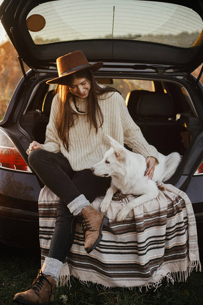 Путешествие и поездка с домашним животным. Стильная женщина в шляпе и свитере сидит и обнимает симпатичную собаку в багажнике машины в вечернем осеннем поле. Молодая хипстерка путешествует с белой собакой. - Фото, изображение