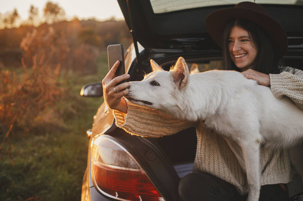 Autumn road trip with pet and travel. Stylish hipster woman taking selfie photo with cute dog in car trunk in sunset light in field. Young female in hat and sweater using phone with sweet white dog - Foto, imagen