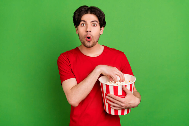 Photo of shocked funky guy hold popcorn box open mouth wear red t-shirt isolated green color background - Foto, immagini