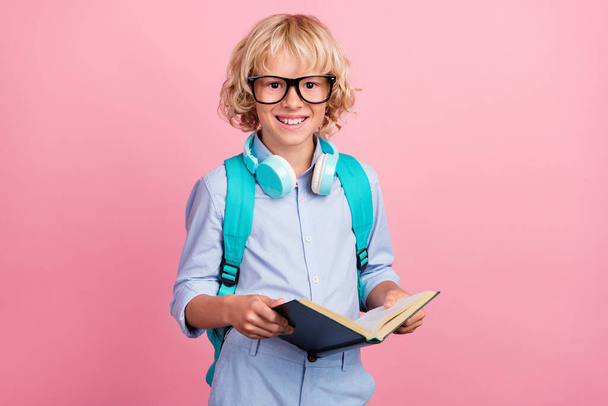 Foto van zoete funky school jongen dragen blauwe shirt bril rugzak oortelefoons glimlachend lezen boek geïsoleerde roze kleur achtergrond - Foto, afbeelding