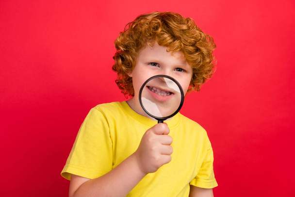 Photo of positive candid ginger small boy magnifying glass show teeth wear yellow t-shirt isolated red color background - Zdjęcie, obraz