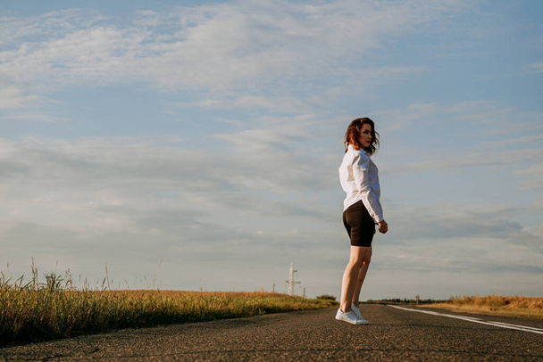 A red-haired woman in a white shirt walks along the road among the fields - Valokuva, kuva