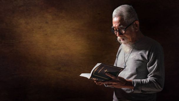 christianity background of senior old christian man with white hair and beard wearing eyeglasses holding and reading holy bible book - Foto, imagen