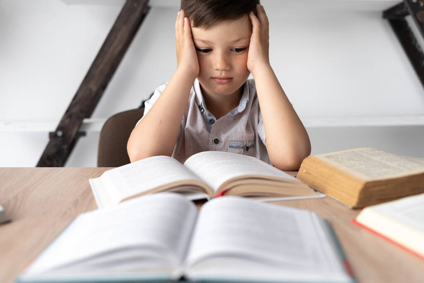 Portrait of upset boy with textbooks and headache. Schoolboy at a desk with open books in the library. Student stress. Self-education concept. High quality photo - Photo, Image