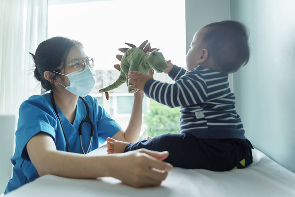 Asian female Pediatrician doctor wearing protective face mask playing dinosaur doll with her little baby boy patient before checkup in medical room at hospital. - Zdjęcie, obraz