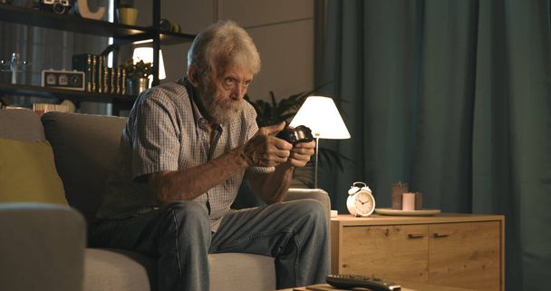 Senior man sitting on the couch at home and playing video games, entertainment and leisure concept - Foto, Imagen