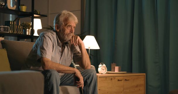 Hombre mayor que se relaja en casa por la noche, está sentado en el sofá y viendo un interesante programa de televisión - Foto, Imagen