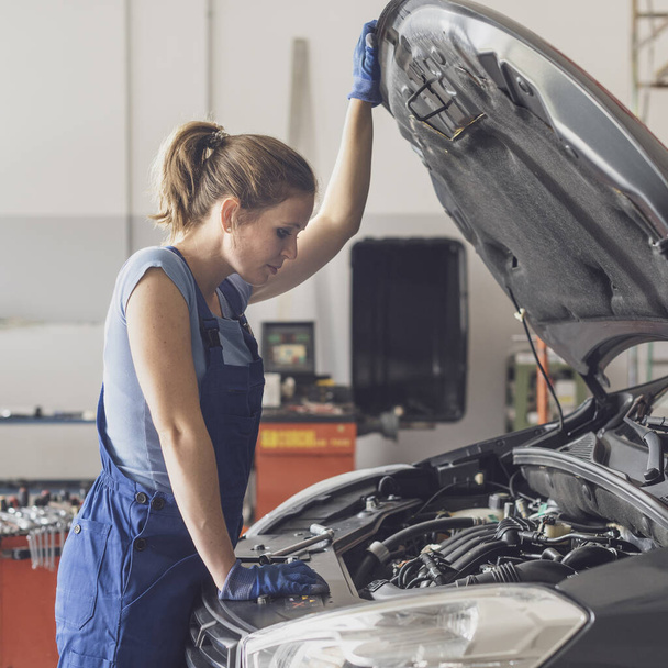 Confident female mechanic working in the auto repair shop, she is checking a car engine - Φωτογραφία, εικόνα