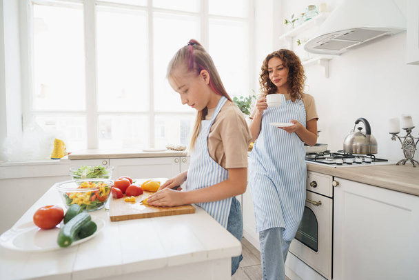 Mommy teaching her teen daughter to cook vegetable salad in kitchen - Foto, imagen