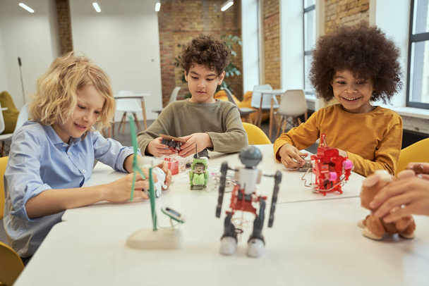 Inovace. Zvědaví malí různorodí chlapci se usmívají, zatímco sedí u stolu a kontrolují robotické hračky, tráví spolu čas ve strojním klubu - Fotografie, Obrázek