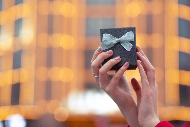 Woman dressed in warm winter coat holding a gift box with a bow at the street with blurred lights. Space for text - Photo, Image