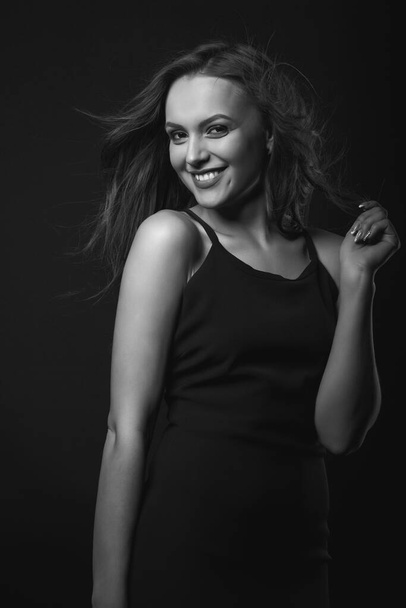 Das Porträt einer glücklichen brünetten Frau mit perfektem Make-up und perfekter Frisur trägt ein klassisches schwarzes Kleid, das in einem dunklen Studio posiert. Schwarz-Weiß-Aufnahmen - Foto, Bild
