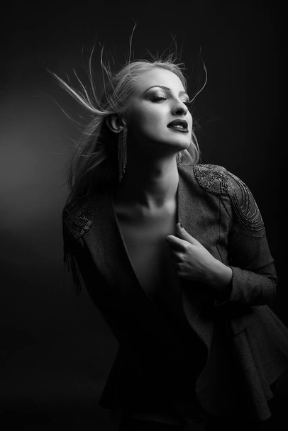 Fashionable young model with bright makeup and long fluttering hair wears jacket. Black and white studio shot - Фото, зображення