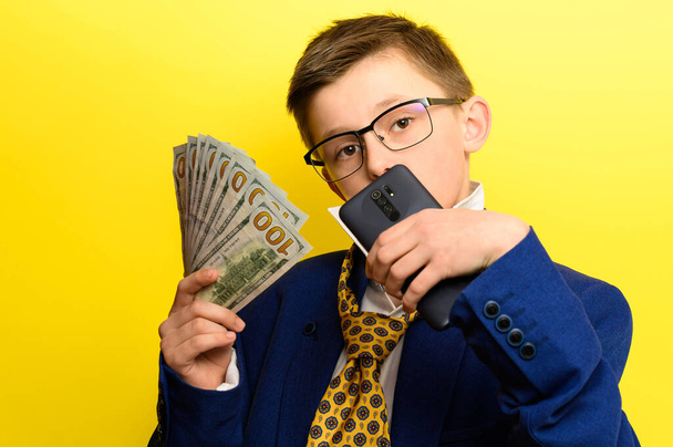 Мальчик в костюме взрослого человека на желтом фоне, международная валюта в руках ребенка, детские сбережения на будущее. новый - Фото, изображение