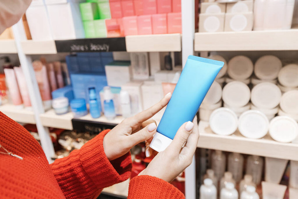 Mujer cliente elige y compra cosméticos en una tienda o farmacia y tiene un tubo de crema hidratante o loción en la mano - Foto, imagen