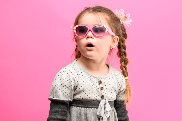 Χαριτωμένο και μικρό κορίτσι σε ροζ φόντο, μακριά κοτσιδάκια και ένα γκρι φόρεμα, ένα κορίτσι με γυαλιά. νέα - Φωτογραφία, εικόνα