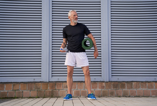 Full length shot του μεσήλικα αθλητή που κάνει αθλητισμό σε εξωτερικούς χώρους, στέκεται με στρώμα γιόγκα - Φωτογραφία, εικόνα