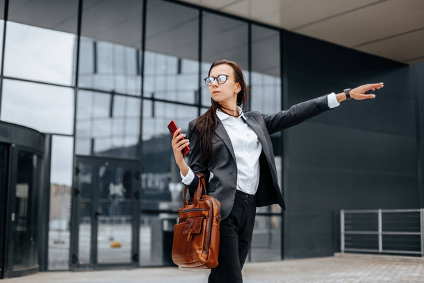 Businesswoman catches a taxi. A woman is waiting for a car and will go to a business meeting. Business concept. Girl with glasses in the city near work. - 写真・画像