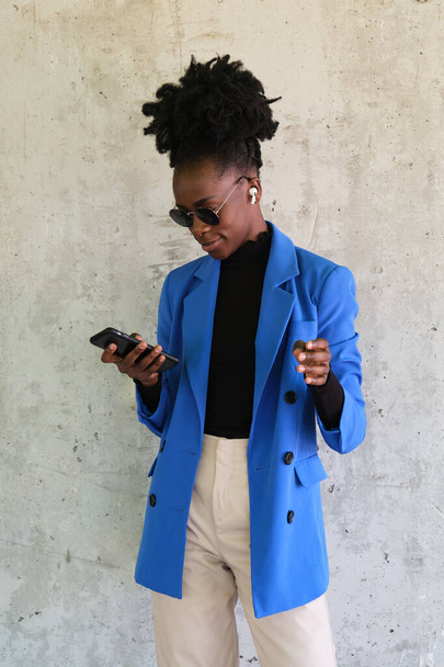 Portrait of smiling young african woman wearing wireless earbuds, selecting the music with her smartphone, wearing blue jacket suit and sunglasses over a concrete wall. - Photo, Image