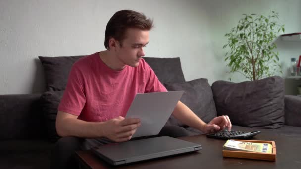 Young guy sitting on the couch by the table with a laptop and counting money for household expenses, the concept of accounting for money at home - Footage, Video