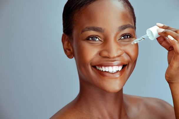 Portrait of african american woman applying liquid cosmetic oil for facial treatment against blue background with copy space. Beauty black middle aged woman with natural makeup holding glass dropper. Close up face of mature lady applying serum around - Photo, image