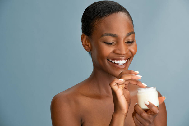 Beautiful mature african woman holding jar of moisturizer. Happy mid black woman holding a bottle of anti-aging lotion isolated over blue background with copy space. Cheerful young black lady applying organic face cream. - Foto, Bild