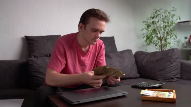 Junger Mann sitzt mit Laptop auf der Couch am Tisch und zählt Geld für Haushaltsausgaben, das Konzept der Abrechnung zu Hause - Filmmaterial, Video