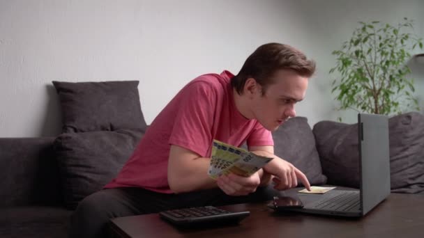 Mladý muž sedí na gauči u stolu s notebookem a počítá peníze na výdaje na domácnost, koncept účtování peněz doma - Záběry, video