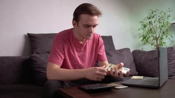 Giovane ragazzo seduto sul divano al tavolo con un computer portatile e contando i soldi per le spese di famiglia, il concetto di contabilità per i soldi a casa - Filmati, video