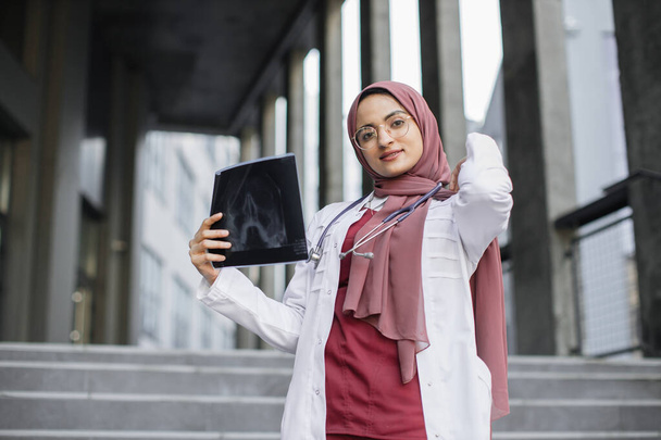 Young Arab woman physician in headscarf holding x-ray, standing outside modern clinic - Foto, imagen