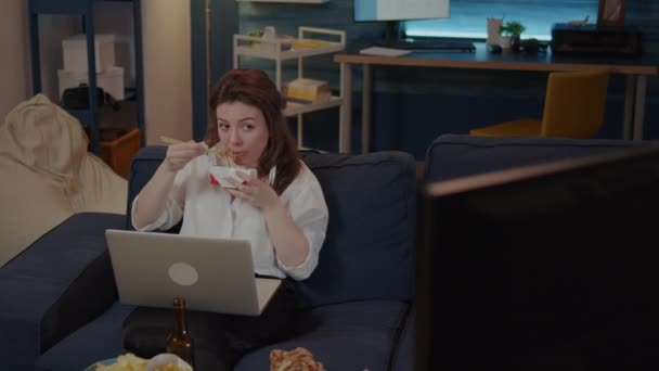 Młoda kobieta korzystająca z laptopa na kanapie podczas jedzenia azjatyckiego jedzenia - Materiał filmowy, wideo