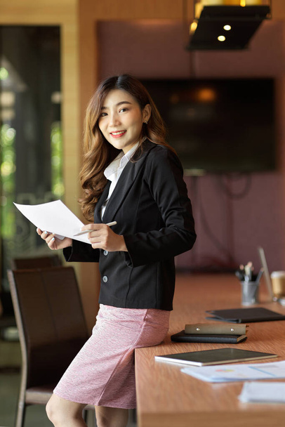 Beautiful female secretary standing and leaning on meeting table, business papers in hands, eyes on camera, meeting room in background - Photo, Image