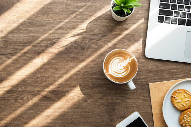 Houten bureautafel met laptop, een schotel amandelkoekjes en een kop koffie met latte 's morgens zonlicht. Werken vanuit huis concept. Bovenaanzicht met kopieerruimte, vlakke lay. - Foto, afbeelding
