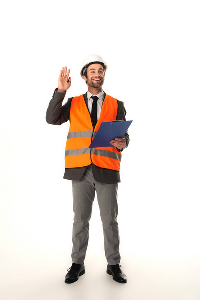 Smiling architect with clipboard waving hand on white background  - Foto, Imagen