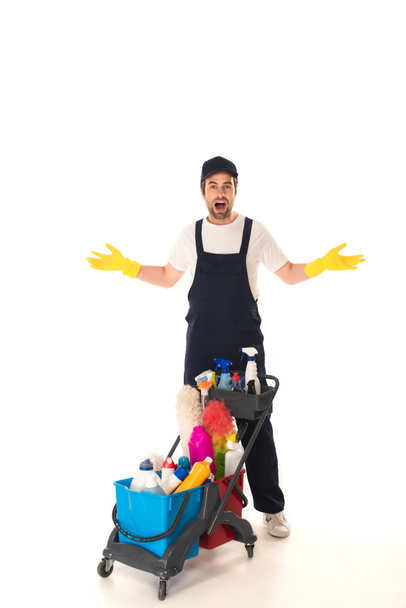 Amazed cleaner in rubber gloves looking at camera near cart on white background  - 写真・画像