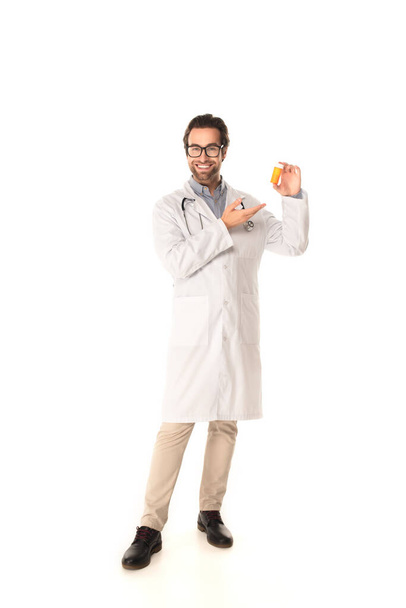 Glimlachende dokter wijzend naar pot met pillen op witte achtergrond  - Foto, afbeelding