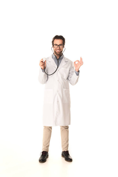 Smiling doctor holding stethoscope and showing okay gesture on white background  - Foto, Imagen